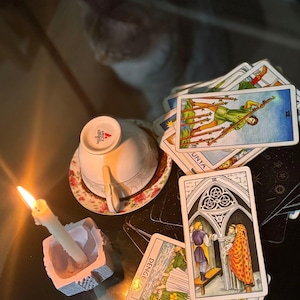 May include: A collection of tarot cards spread out on a surface, including the Seven of Wands, Three of Pentacles, and Temperance cards. A white teacup sits upside down on a floral saucer next to a lit white candle in a square holder.