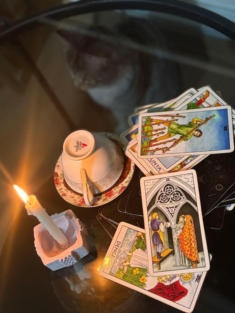 May include: A collection of tarot cards spread out on a surface, including the Seven of Wands, Three of Pentacles, and Temperance cards. A white teacup sits upside down on a floral saucer next to a lit white candle in a square holder.