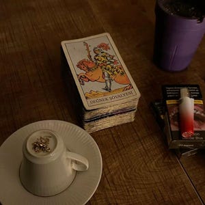 May include: A deck of tarot cards featuring the Knight of Wands, labeled DEĞNEK Ş&Ouml;VALYESI, sits next to a white teacup turned upside down on a saucer, a purple ashtray, and a pack of cigarettes with a red and white lighter on top.