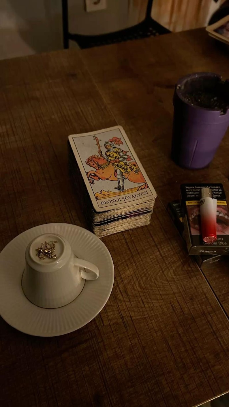 May include: A deck of tarot cards featuring the Knight of Wands, labeled DEĞNEK Ş&Ouml;VALYESI, sits next to a white teacup turned upside down on a saucer, a purple ashtray, and a pack of cigarettes with a red and white lighter on top.