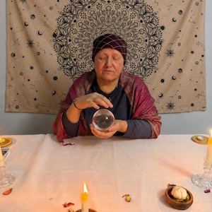 May include: A clear glass crystal ball held by a person wearing a dark grey long-sleeved shirt, a maroon patterned shawl, and a dark maroon head covering with a white grid pattern, positioned before a beige tapestry featuring a black mandala design.