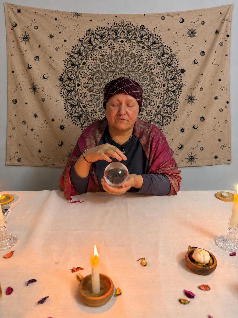 May include: A clear glass crystal ball held by a person wearing a dark grey long-sleeved shirt, a maroon patterned shawl, and a dark maroon head covering with a white grid pattern, positioned before a beige tapestry featuring a black mandala design.