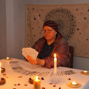 May include: A person wearing a dark headscarf and shawl holds a fan of tarot cards over a white tablecloth. The table is decorated with multiple lit candles in wooden and glass holders, scattered flower petals, and a spread of tarot cards.