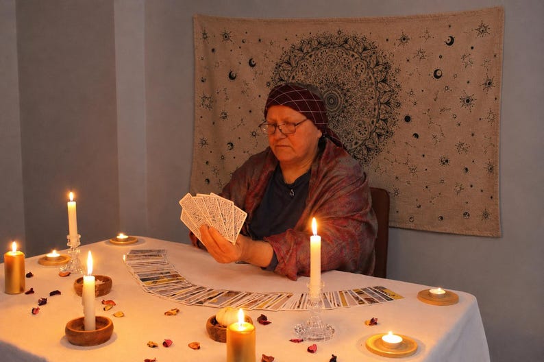 May include: A person wearing a dark headscarf and shawl holds a fan of tarot cards over a white tablecloth. The table is decorated with multiple lit candles in wooden and glass holders, scattered flower petals, and a spread of tarot cards.