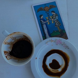 May include: A white ceramic coffee cup containing dark coffee grounds and a matching saucer with a circular coffee stain, positioned next to a Two of Cups tarot card featuring two figures holding golden chalices beneath a winged lion symbol.