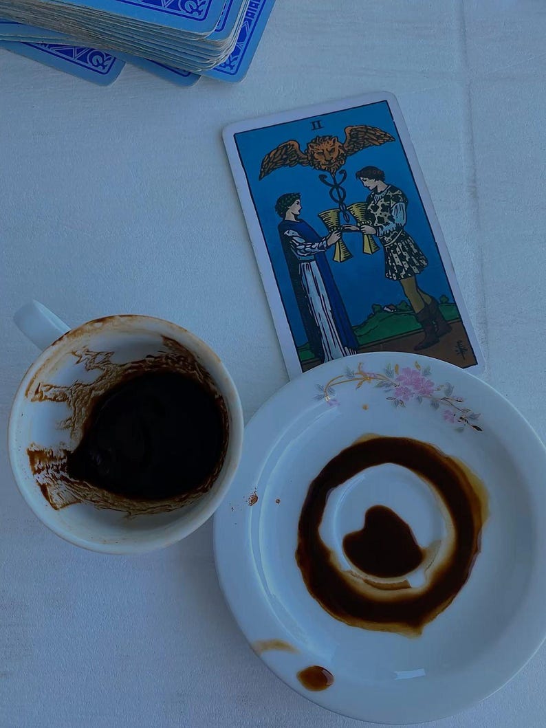 May include: A white ceramic coffee cup containing dark coffee grounds and a matching saucer with a circular coffee stain, positioned next to a Two of Cups tarot card featuring two figures holding golden chalices beneath a winged lion symbol.