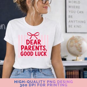 Könnte beinhalten: Ein weißes kurzärmeliges T-Shirt mit einem roten Grafikdruck einer Schleife über vertikalen hellrosa Streifen und dem roten Text "DEAR PARENTS, GOOD LUCK". Das Design ist mittig auf der Brust platziert und wird mit einer blauen Jeans kombiniert.