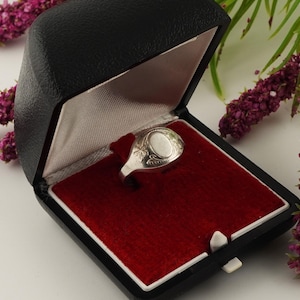 May include: A silver signet ring featuring an oval face with engraved floral detailing on the shoulders. The ring is displayed inside a black hinged jewellery box with a red velvet interior lining and a white satin lid lining.