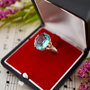 May include: A ring featuring a large, round, faceted light blue gemstone set in a metallic band with decorative prongs, presented inside a black jewelry box with a vibrant red velvet interior lining and a white satin lid lining.