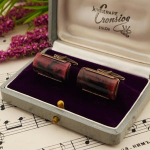 Peut inclure: Une paire de boutons de manchette rectangulaires au motif marbré rouge et noir, montés sur un cadre métallique. Ils sont présentés dans un coffret gris avec un intérieur en velours violet et l'inscription Juvelerare Cronsioe Eslöv à l'intérieur.