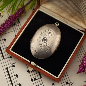 May include: An oval-shaped silver-toned metal locket featuring an engraved floral pattern on the front surface and a single clear faceted stone set in the centre. The locket includes a small metal loop at the top for attachment to a chain.
