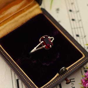 Può includere: Un anello in argento con una pietra preziosa rettangolare rosso scuro sfaccettata, fissata con una montatura a quattro griffe. Il gambo è sottile e si divide in due bracci delicati verso la pietra, conferendo un aspetto raffinato al gioiello.