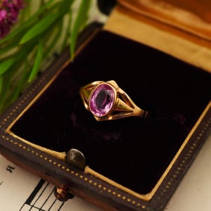 Op de afbeelding: Een gouden ring met een centrale ovaal geslepen roze edelsteen. De ringband heeft een gespleten ontwerp dat taps toeloopt naar de zetting. De ring wordt gepresenteerd in een donkerbruine rechthoekige doos met een paarse fluwelen voering.