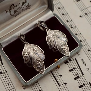 May include: A pair of silver-toned drop earrings featuring an intricate leaf-shaped design with embossed floral patterns and dark oxidized detailing. The earrings are presented in a grey hinged jewelry box with a dark purple velvet interior.