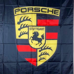 May include: A black rectangular flag featuring the Porsche logo in the center. The logo displays a yellow shield with black antlers, red stripes, and a central crest with a black horse and the text STUTTGART. The word PORSCHE appears in white.