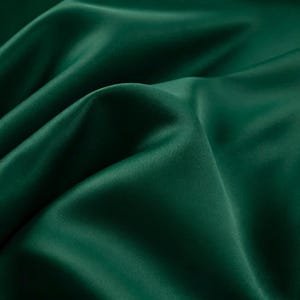 May include: A deep forest green fabric with a smooth, lustrous finish. The material is draped in soft, flowing folds that create subtle highlights and shadows across the surface, emphasizing the rich, saturated color and the silky texture.