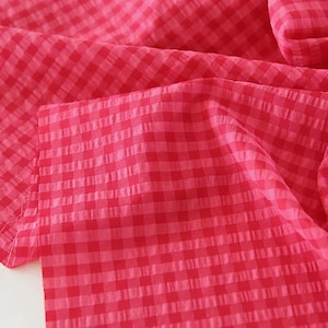 May include: A vibrant red and white gingham fabric featuring a classic checkered pattern. The material has a distinct seersucker texture with a puckered surface, creating a lightweight and slightly crinkled appearance across the folded textile.