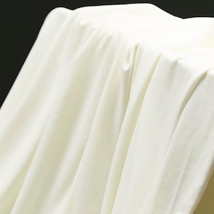 May include: A solid off-white fabric draped in soft, vertical folds. The material has a smooth, matte texture and a consistent light cream color throughout, creating deep shadows within the creases of the fabric as it hangs downward.