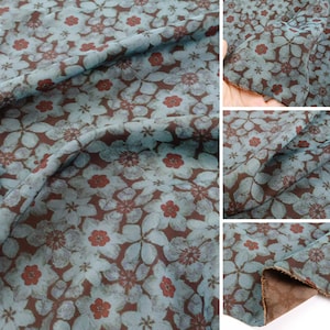 May include: A fabric featuring a repeating floral pattern with light blue petals and small dark red centers set against a dark brown background. The material has a soft, draped texture with visible folds and a finished edge showing the brown underside.