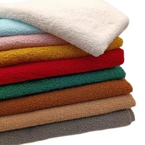 May include: A stack of folded sherpa fleece fabric in various colours including white, light blue, light pink, mustard yellow, red, dark green, brown, tan, and grey. Each piece of fabric features a thick, textured, and curly pile surface.