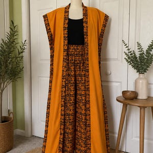 May include: A sleeveless, floor-length mustard yellow duster coat featuring wide panels of a black and orange textured abstract print along the front edges and armholes, paired with matching wide-leg trousers and a solid black tank top.