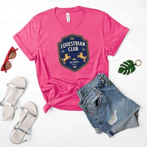 Puede incluir: Una camiseta rosa brillante de manga corta con un gráfico de escudo azul oscuro con el texto The Equestrian Club, Mares Only y dos siluetas de caballos doradas. Incluye pantalones cortos de mezclilla azules, sandalias plateadas, gafas de sol rojas y un llavero de hoja verde.