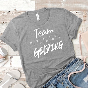 Puede incluir: Una camiseta gris jaspeado de manga corta con el texto blanco Team GELDING impreso en la parte delantera, acompañado de una serie de pequeños iconos de herradura blancos dispuestos en arco entre las dos palabras del texto.