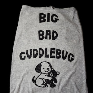 May include: A heather gray sleeveless pet shirt featuring the black glittery text BIG BAD CUDDLEBUG printed in the center. Below the text is a graphic of a puppy hugging a teddy bear, also rendered in black glittery material on the fabric.