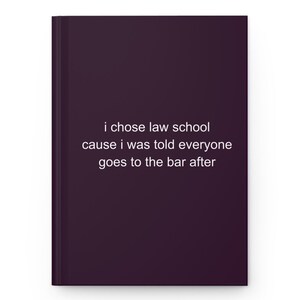 May include: A dark purple hardcover notebook featuring white text centered on the front cover that reads: i chose law school cause i was told everyone goes to the bar after. The notebook has a clean, solid-colored finish and a vertical spine.