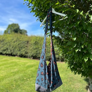 Puede incluir: Un bolso tote reversible con estampado de leopardo en verde azulado y naranja, colgado de una percha blanca en una rama de árbol. El bolso tiene una base gris y un diseño reversible con un estampado de rayas rojas y blancas en el interior. El fondo muestra hierba verde y cielo azul.