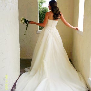 Peut inclure: Une robe de mari&eacute;e ivoire sans bretelles, longue jusqu'au sol, avec un corsage ajust&eacute; et une jupe ample et fluide qui se prolonge en tra&icirc;ne. La robe pr&eacute;sente un d&eacute;tail de taille en forme de V subtil et une superposition de tulle. Un bouquet est tenu.
