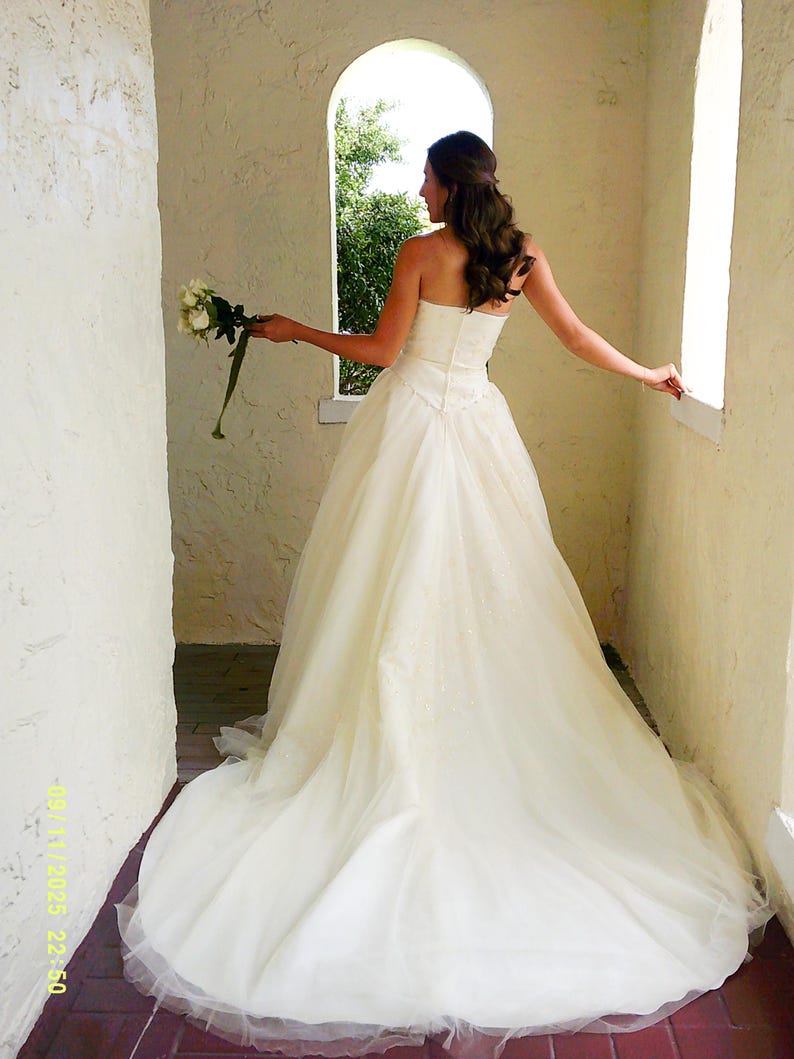 Peut inclure: Une robe de mari&eacute;e ivoire sans bretelles, longue jusqu'au sol, avec un corsage ajust&eacute; et une jupe ample et fluide qui se prolonge en tra&icirc;ne. La robe pr&eacute;sente un d&eacute;tail de taille en forme de V subtil et une superposition de tulle. Un bouquet est tenu.