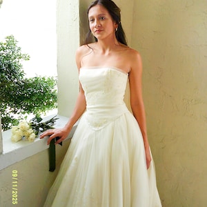 Peut inclure: Une robe de mari&eacute;e bustier ivoire dot&eacute;e d'un corsage ajust&eacute; avec de subtiles broderies florales et d'une jupe en tulle longue et volumineuse. La robe pr&eacute;sente une taille structur&eacute;e et des ornements d&eacute;licats. Un bouquet de roses blanches est pos&eacute;.