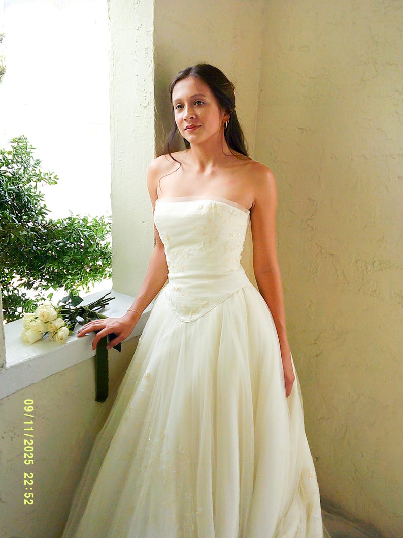 Peut inclure: Une robe de mari&eacute;e bustier ivoire dot&eacute;e d'un corsage ajust&eacute; avec de subtiles broderies florales et d'une jupe en tulle longue et volumineuse. La robe pr&eacute;sente une taille structur&eacute;e et des ornements d&eacute;licats. Un bouquet de roses blanches est pos&eacute;.