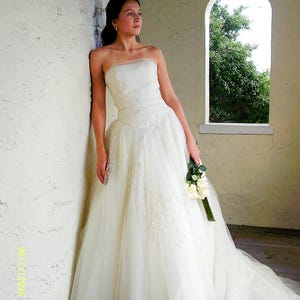 Peut inclure: Une robe de mari&eacute;e ivoire sans bretelles, longue jusqu'au sol, dot&eacute;e d'un corsage ajust&eacute; avec des broderies florales d&eacute;licates et des perles, se prolongeant par une jupe en tulle volumineuse et superpos&eacute;e avec une longue tra&icirc;ne, tenant un bouquet.