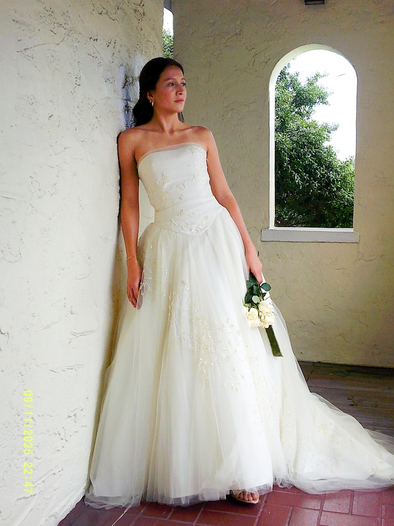 Peut inclure: Une robe de mari&eacute;e ivoire sans bretelles, longue jusqu'au sol, dot&eacute;e d'un corsage ajust&eacute; avec des broderies florales d&eacute;licates et des perles, se prolongeant par une jupe en tulle volumineuse et superpos&eacute;e avec une longue tra&icirc;ne, tenant un bouquet.