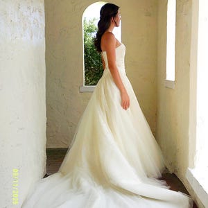 Peut inclure: Une robe de mari&eacute;e bustier couleur ivoire avec un corsage ajust&eacute; et une jupe volumineuse en tulle arrivant au sol, dot&eacute;e d'une longue tra&icirc;ne. La robe est compos&eacute;e de plusieurs couches de tissu transparent cr&eacute;ant une silhouette ample et &eacute;tendue.
