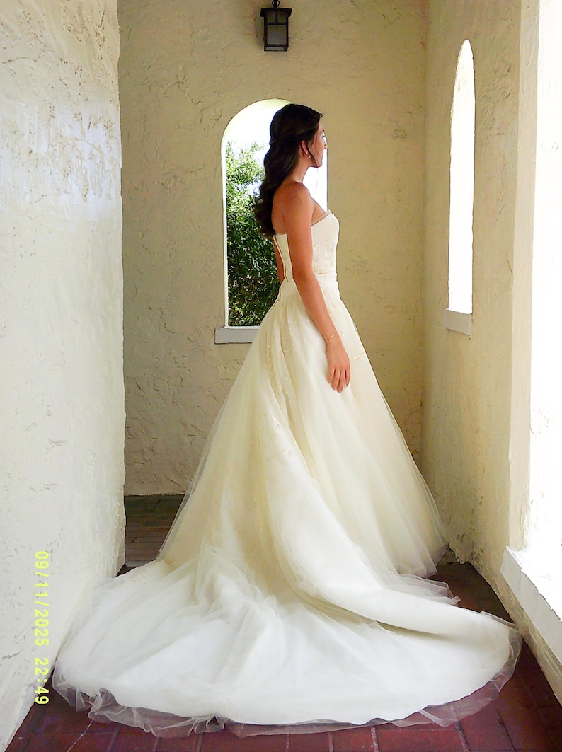 Peut inclure: Une robe de mari&eacute;e bustier couleur ivoire avec un corsage ajust&eacute; et une jupe volumineuse en tulle arrivant au sol, dot&eacute;e d'une longue tra&icirc;ne. La robe est compos&eacute;e de plusieurs couches de tissu transparent cr&eacute;ant une silhouette ample et &eacute;tendue.