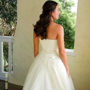 Peut inclure: Une robe ivoire sans bretelles arrivant au sol, dot&eacute;e d'un corsage ajust&eacute; avec une fermeture &eacute;clair verticale centrale et d'une jupe en tulle superpos&eacute;e. Le corsage et la jupe sont orn&eacute;s de broderies florales discr&egrave;tes et de petites perles.