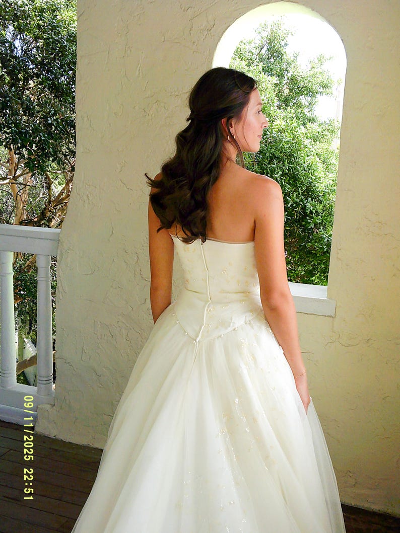 Peut inclure: Une robe ivoire sans bretelles arrivant au sol, dot&eacute;e d'un corsage ajust&eacute; avec une fermeture &eacute;clair verticale centrale et d'une jupe en tulle superpos&eacute;e. Le corsage et la jupe sont orn&eacute;s de broderies florales discr&egrave;tes et de petites perles.