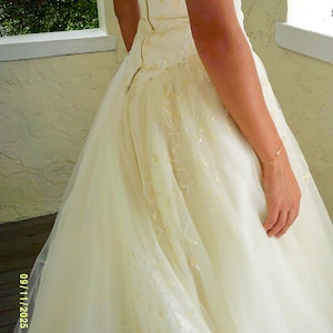 Peut inclure: Une robe longue couleur blanc cass&eacute; dot&eacute;e d'un corsage ajust&eacute; avec une rang&eacute;e verticale de petits boutons sur le c&ocirc;t&eacute; et une jupe en tulle superpos&eacute;e, orn&eacute;e de d&eacute;licates broderies florales ton sur ton et de perles subtiles sur tout le tissu.