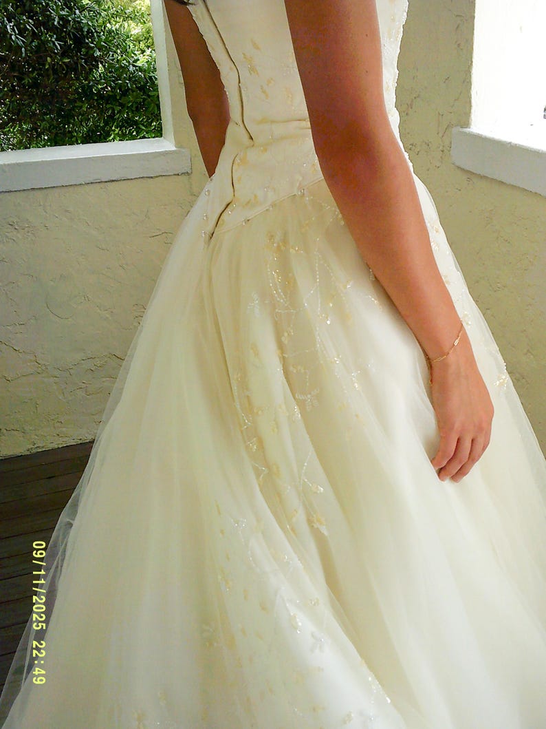 Peut inclure: Une robe longue couleur blanc cass&eacute; dot&eacute;e d'un corsage ajust&eacute; avec une rang&eacute;e verticale de petits boutons sur le c&ocirc;t&eacute; et une jupe en tulle superpos&eacute;e, orn&eacute;e de d&eacute;licates broderies florales ton sur ton et de perles subtiles sur tout le tissu.
