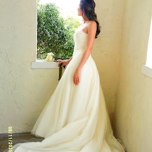 Peut inclure: Une robe de mari&eacute;e ivoire sans bretelles, longue jusqu'au sol, avec un corsage ajust&eacute; et une jupe volumineuse en tulle superpos&eacute; qui se prolonge en une longue tra&icirc;ne. La robe pr&eacute;sente des d&eacute;tails textur&eacute;s subtils sur le corsage.