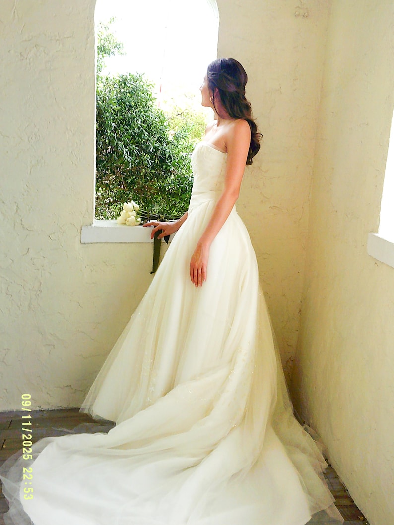 Peut inclure: Une robe de mari&eacute;e ivoire sans bretelles, longue jusqu'au sol, avec un corsage ajust&eacute; et une jupe volumineuse en tulle superpos&eacute; qui se prolonge en une longue tra&icirc;ne. La robe pr&eacute;sente des d&eacute;tails textur&eacute;s subtils sur le corsage.