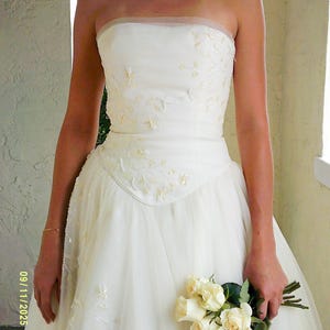 Peut inclure: Une robe bustier ivoire dot&eacute;e d'un corsage ajust&eacute; avec des perles florales d&eacute;licates et des sequins. La taille est en forme de V, se prolongeant par une jupe en tulle superpos&eacute;e. Un bouquet de roses blanches est tenu, avec la date 09/11/2025 22:42 visible.