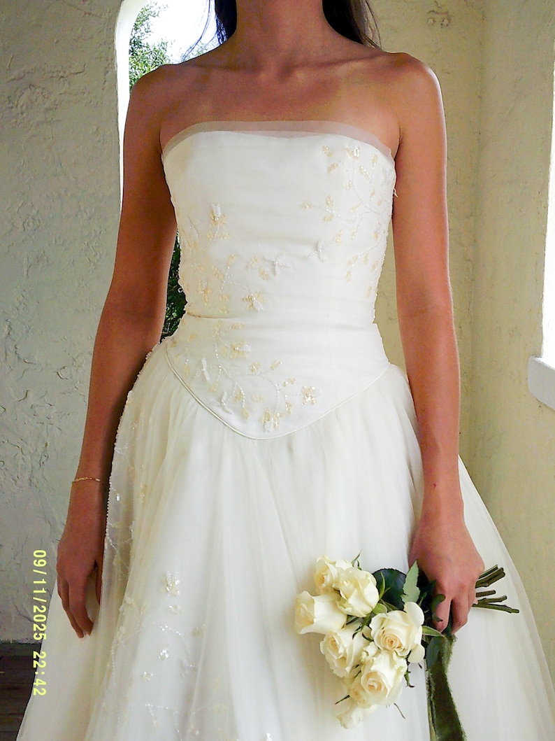 Peut inclure: Une robe bustier ivoire dot&eacute;e d'un corsage ajust&eacute; avec des perles florales d&eacute;licates et des sequins. La taille est en forme de V, se prolongeant par une jupe en tulle superpos&eacute;e. Un bouquet de roses blanches est tenu, avec la date 09/11/2025 22:42 visible.