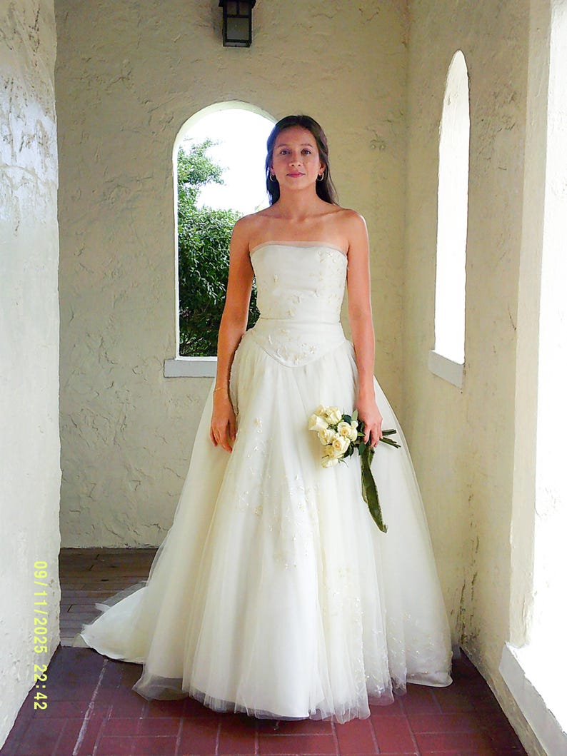 Peut inclure: Une robe de mari&eacute;e ivoire sans bretelles, longue jusqu'au sol, dot&eacute;e d'un corsage ajust&eacute; avec une broderie subtile et d'une jupe en tulle volumineuse &agrave; plusieurs &eacute;paisseurs. La robe est accompagn&eacute;e d'un petit bouquet de roses cr&egrave;me.