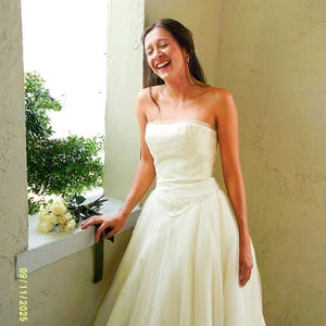 Peut inclure: Une robe de mari&eacute;e ivoire sans bretelles, longue jusqu'au sol, dot&eacute;e d'un corsage ajust&eacute; avec une broderie subtile et d'une jupe en tulle superpos&eacute;e. La robe pr&eacute;sente une encolure droite et une taille marqu&eacute;e, avec un bouquet de roses blanches.