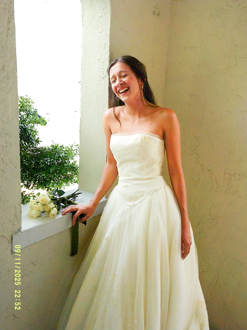 Peut inclure: Une robe de mari&eacute;e ivoire sans bretelles, longue jusqu'au sol, dot&eacute;e d'un corsage ajust&eacute; avec une broderie subtile et d'une jupe en tulle superpos&eacute;e. La robe pr&eacute;sente une encolure droite et une taille marqu&eacute;e, avec un bouquet de roses blanches.