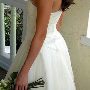 Peut inclure: Une robe bustier ivoire avec un corsage ajust&eacute; orn&eacute; de perles et de sequins d&eacute;licats, se prolongeant par une jupe en tulle superpos&eacute;e. Un fin cordon de petites perles blanches souligne la taille, accompagn&eacute; d'un bouquet de roses blanches.